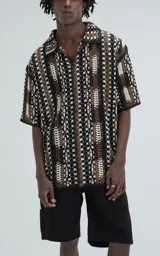Camisa de manga corta con tejido de punto abierto tipo crochet, corte oversize y cuello camisero. Presenta un diseño de rayas verticales con patrón geométrico en tonos negro, marrón y blanco.