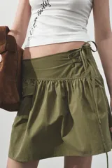 Falda corta de tiro medio en color verde militar. Presenta un diseño con volumen, pliegues, un bolsillo de plastrón en el delantero y detalle de trabilla con hebilla metálica en la cintura. Incluye cierre lateral con cremallera oculta.