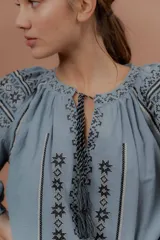 Blusa de estilo bohemio color celeste, con mangas abullonadas y amplias, escote redondo con tiras colgantes y borlas, y detalles de bordados geométricos en contraste de color en el pecho y las mangas.