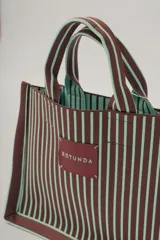 Bolso tote marrón con rayas verticales verde agua. Tiene doble asa corta marrón con rayas verde agua y etiqueta cuadrada con el nombre de la marca en el frente.
