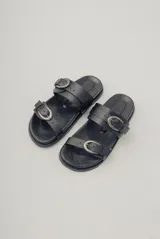 Sandalias planas de cuero negro con diseño de dos tiras ajustables con hebillas metálicas.