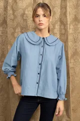 Camisa de jean azul con cuello babero, detalles de volados y bordados en contraste. Presenta mangas largas con puños elásticos y cierre frontal con botones.