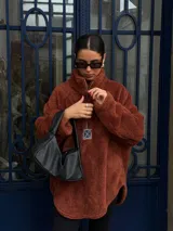 Buzo de corderito color marrón, con cuello alto y cierre frontal parcial. Presenta un diseño holgado con mangas largas y un parche decorativo en el frente.