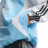 Campera deportiva Adidas de la Selección Argentina, modelo retro inspirado en el diseño de 2006. Presenta un diseño en blanco y celeste con líneas curvas, cuello alto, cierre frontal y las icónicas tres tiras negras en las mangas.