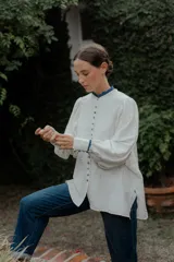 Camisa de corte oversize color crudo, con cuello mao y puños con detalle de volados en color azul. Presenta cierre frontal con botones forrados y mangas largas abullonadas.