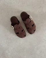 Zueco tipo sandalia de estilo jaula, confeccionado en gamuza negra con tiras entrelazadas y hebilla metálica dorada lateral. Cuenta con plantilla acolchada con logo de la marca.