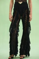 Mono strapless negro confeccionado en encaje con diseño calado. Presenta un corpiño con detalle de frunce y hebilla dorada en el centro, cintura marcada con cinturón fino y hebilla dorada, y piernas acampanadas con volados laterales de encaje.