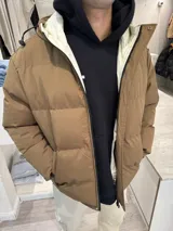 Campera puffer de color marrón claro con diseño acolchado, cierre frontal completo y capucha ajustable con cordones. Presenta un forro interno en color crema y bolsillos laterales.