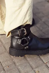 Bota corta negra de cuero sintético con suela track, caña baja y correas con hebillas metálicas.