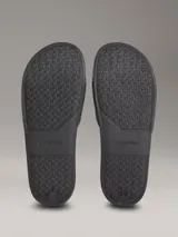 Ojotas tipo slide de color negro con tira superior de piel sintética que presenta un patrón grabado del logo de Calvin Klein. Poseen plantilla ergonómica moldeada y suela de EVA ligera.