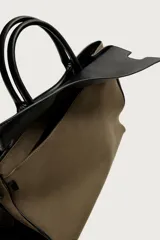 Bolso shopper de mano combinando cuerpo de gamuza color verde militar con solapa superior, asas y detalles en cuero sintético negro. Cuenta con compartimento principal con cierre imantado, bolsillo interior con cierre y bolsillo frontal plano con solapa.