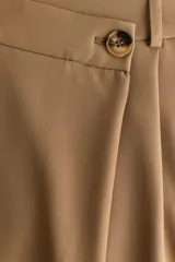 Pantalón sastrero de corte holgado en color tostado, con diseño asimétrico cruzado en la cintura, botón frontal y pliegues en las piernas.