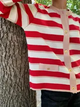 Chaqueta de punto color rojo y blanco a rayas horizontales, con ribetes y bolsillos en color rosa pálido. Presenta cierre frontal con botones dorados.