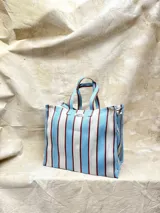 Bolso tipo tote grande con estampado a rayas verticales en tonos celeste, blanco y bordó. Tiene doble asa corta de mano y cierre superior.