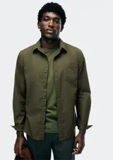 Camisa de manga larga color verde oliva, con cuello clásico y un bolsillo de parche en el pecho izquierdo. Se lleva abierta sobre una remera verde más clara.