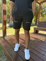 Bermuda deportiva negra de felpa, con bolsillos y cordón ajustable en la cintura. Presenta el texto "TOP STYLE SINCE 2021" estampado en blanco en la pierna derecha y "follow your dreams" en cursiva en la pierna izquierda.