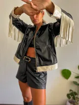 Campera negra de cuero sintético con cierre frontal, detalles en blanco en cuello, puños y ruedo, y flecos blancos en las mangas.