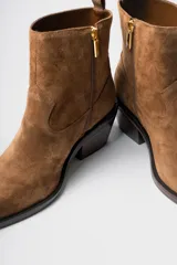 Bota corta de estilo western confeccionada en cuero gamuzado color marrón. Presenta un diseño de caña media con tirador trasero, corte asimétrico en el empeine, punta cuadrada y taco bloque de madera.