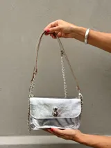 Minibag de cuero metalizado plateado con diseño tipo acordeón. Cuenta con una solapa frontal con cierre metálico a presión y una correa combinada de cadena metálica y cuero.