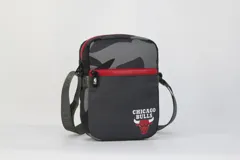 Morral bandolera negro con estampado camuflado en tonos grises en la parte superior y logo de los Chicago Bulls bordado en rojo y blanco en la parte inferior. Tiene doble cierre rojo, correa regulable y logo de la NBA en el lateral.