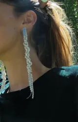 Aros colgantes largos con diseño trenzado y detalles de strass brillante. Cuentan con una base rectangular metálica en la parte superior y terminaciones en tiras finas de cristales.