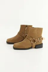 Bota de caña media confeccionada en cuero con acabado de gamuza en color mostaza. Presenta una correa decorativa desmontable con argolla y hebilla metálica dorada, cierre lateral interno para facilitar el calce y suela de goma negra con taco bajo cuadrado.