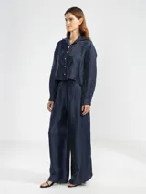 Camisa de manga larga color azul marino, confeccionada en lino italiano con hilo metalizado que le da un sutil brillo. Presenta cuello clásico y botones frontales, con un detalle de broche o dije en forma de flor en el cuello.