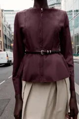 Chaqueta color beige con efecto ante, cuello redondo, manga larga y cierre frontal con cremallera. Presenta un bajo en evasé y un cinturón fino de cuero marrón oscuro con hebilla metálica.