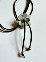 Gargantilla con dije de flor plateada con strass en el centro y tiras de gamuza negra con terminales plateadas.