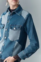 Campera recta de denim rígido con combinación de recortes en tonos de azul. Cuenta con bolsillos de solapa en el pecho y botones metálicos. Las mangas tienen puños con encaje negro.