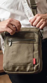 Morral de tela canvas color verde militar con detalles en cuero sintético. Presenta un compartimento principal con cierre superior, dos bolsillos frontales con cremallera y un bolsillo trasero. Incluye una correa de hombro ajustable y etiqueta de marca lateral.