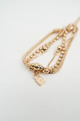 Brazalete multicapa de color dorado compuesto por cadenas de eslabones tipo caja y cadenas de esferas pequeñas, con un dije en forma de cubo en el extremo.