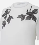 Remera blanca de manga corta y cuello redondo, confeccionada en algodón con bordados florales en tono oscuro aplicados en los hombros.