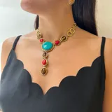 Collar corbatero de bronce con piedras naturales engarzadas. El diseño central presenta una piedra turquesa ovalada, flanqueada por piedras rojas y piedras ojo de tigre ovaladas. De esta piedra central cuelga una cascada de piedras rojas y ojo de tigre engarzadas en marcos de bronce.