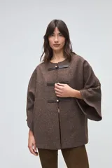 Capa de lana fina sin teñir, con diseño de corte amplio y mangas tipo kimono. Presenta cierre frontal mediante tres alamares de cuero en tono marrón.