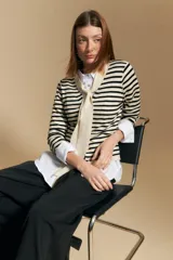 Cárdigan de tejido de punto a rayas horizontales beige y blanco, con escote en V y un lazo ancho anudado al frente. Se lleva sobre una camisa blanca de cuello abotonado con puños anchos.