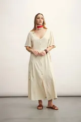 Vestido largo color beige claro, con escote en V, manga corta acampanada y cierre frontal de botones. Presenta un lazo ajustable en la cintura y un tejido con textura sutil.