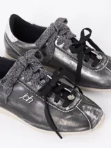 Zapatilla de cuero vacuno en tono gris oscuro metalizado, con detalles en cuero descarne. Presenta doble cordón, uno de ellos tejido, y charms decorativos. Cuenta con un detalle en color plateado en el talón y suela de goma en tono claro.