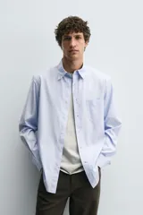 Camisa de algodón de corte regular, color celeste claro con finas rayas blancas verticales. Presenta cuello abotonado (button-down), manga larga con puños abotonados y un bolsillo plastrón en el pecho izquierdo.