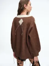 Sweater de tejido de punto con diseño de rombos (argyle) en tonos marrón, verde oliva y crema. Presenta escote en V, mangas largas con puños ajustados y detalles de hilos colgantes decorativos en el frente que le otorgan un aspecto desgastado y moderno.