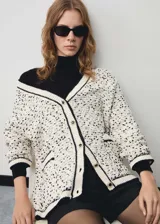 Cardigan de punto grueso con diseño jaspeado en blanco y negro. Presenta cuello en pico, cierre frontal con botones efecto joya, dos bolsillos de parche delanteros y detalles en contraste negro en los bordes.