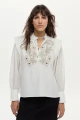 Blusa blanca de tela liviana con bordado detallado en el frente, cuello alto con volado y escote en V con botones. Tiene mangas largas abullonadas con puños amplios y detalles estructurados en los hombros.