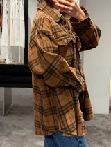 Camisa tipo sobrecamisa de corte oversize, confeccionada en tela a cuadros en tonos marrones y negros. Presenta cierre frontal con botones metálicos a presión, bolsillos frontales con solapa y mangas largas con puños abotonados.