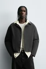 Sobrecamisa negra de algodón con estructura canvas, corte cropped y calce holgado. Presenta cuello solapa, manga larga, bolsillos laterales en cadera y cierre frontal con botones a presión metálicos.