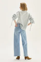 Camisa a rayas verticales de calce recto, con cuello clásico y mangas abullonadas con detalles de frunces y lazos en los puños. Presenta un pequeño bordado en el pecho.