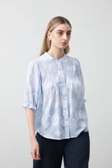 Camisa de algodón con fondo azul claro a rayas finas y estampa floral blanca en relieve. Presenta cuello cerrado, cartera de botones frontal y mangas cortas con volumen y puño elastizado. Posee frunces en el frente que aportan caída.