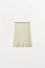 Falda midi de tejido ligero color crema o beige muy claro, con cintura elástica de color beige más oscuro. El bajo está adornado con una aplicación de encaje floral a tono.