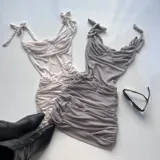Vestido corto de tela fruncida, color gris claro, con escote corazón y tirantes finos anudados en los hombros. Presenta un efecto drapeado en todo el cuerpo.