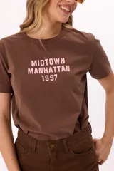 Remera de algodón color marrón con cuello redondo y mangas cortas. Presenta un estampado frontal en color rosado con el texto 'MIDTOWN MANHATTAN 1997' en estilo tipográfico varsity.