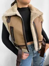 Chaleco color beige con interior de piel sintética color crema, cierre frontal con cremallera, bolsillos laterales con cremallera, cuello con solapas y cinturón ajustable en la cintura.